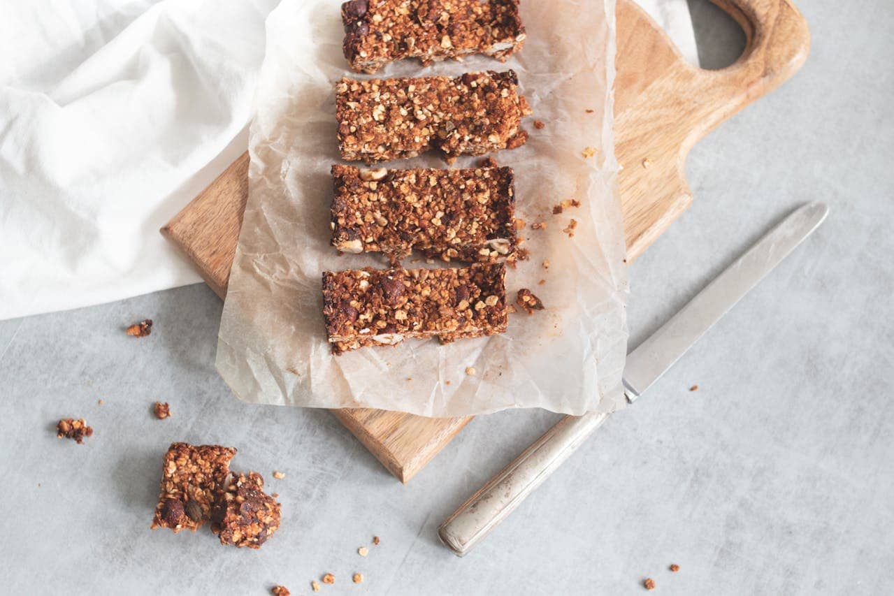 Four homemade granola bars arranged in a row on parchment paper atop a wooden cutting board, with a knife nearby, some crumbs scattered on the light gray surface