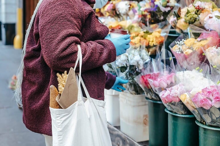 A person wearing a burgundy coat and blue gloves holding a coffee cup, standing near a flower stand, carrying a white tote bag with a baguette and dried wheat, browsing bouquets of colorful flowers wrapped in plastic