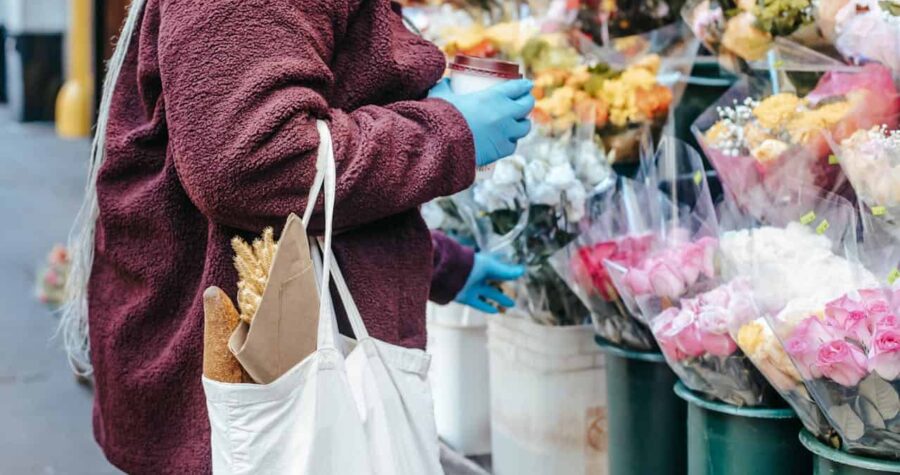 A person wearing a burgundy coat and blue gloves holding a coffee cup, standing near a flower stand, carrying a white tote bag with a baguette and dried wheat, browsing bouquets of colorful flowers wrapped in plastic