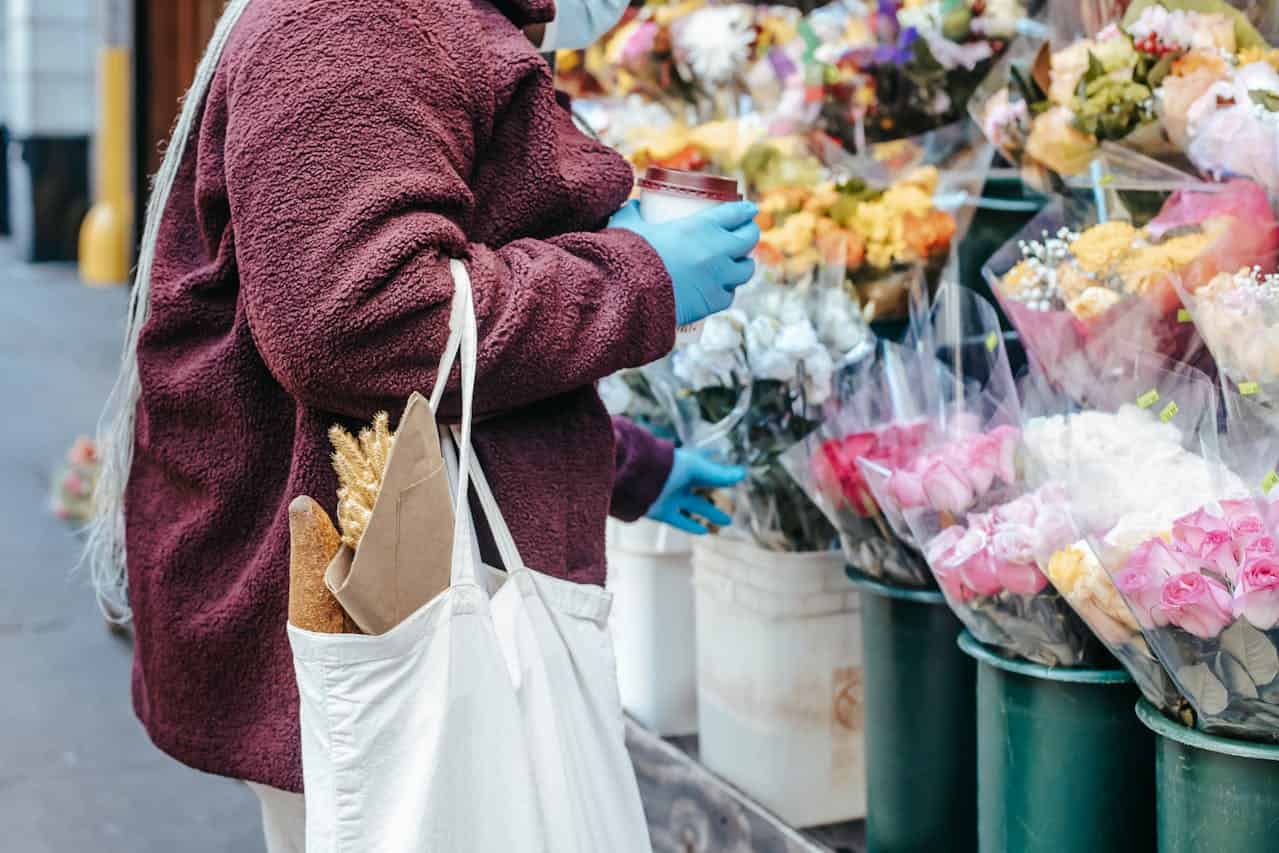 A person wearing a burgundy coat and blue gloves holding a coffee cup, standing near a flower stand, carrying a white tote bag with a baguette and dried wheat, browsing bouquets of colorful flowers wrapped in plastic