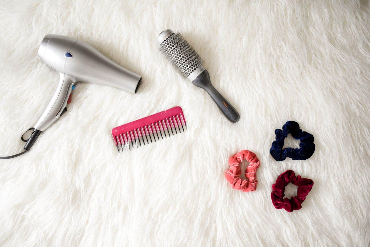 Hair styling tools arranged on white fur background, including silver hair dryer, round brush, pink comb, and three fabric scrunchies in navy, coral, and burgundy colors