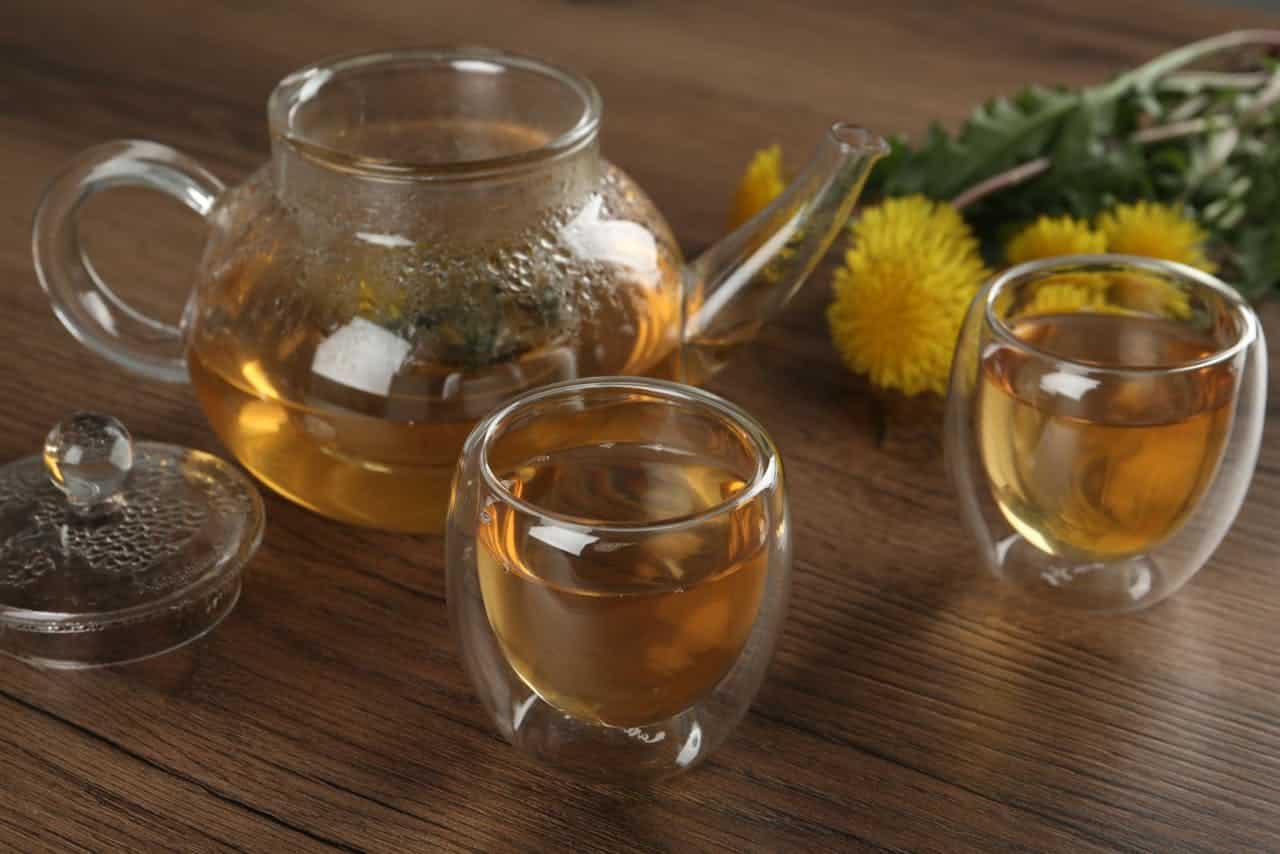 Glass teapot with herbal tea, two double-walled glass cups filled with tea, fresh dandelion flowers and leaves on a wooden table, steam on teapot lid