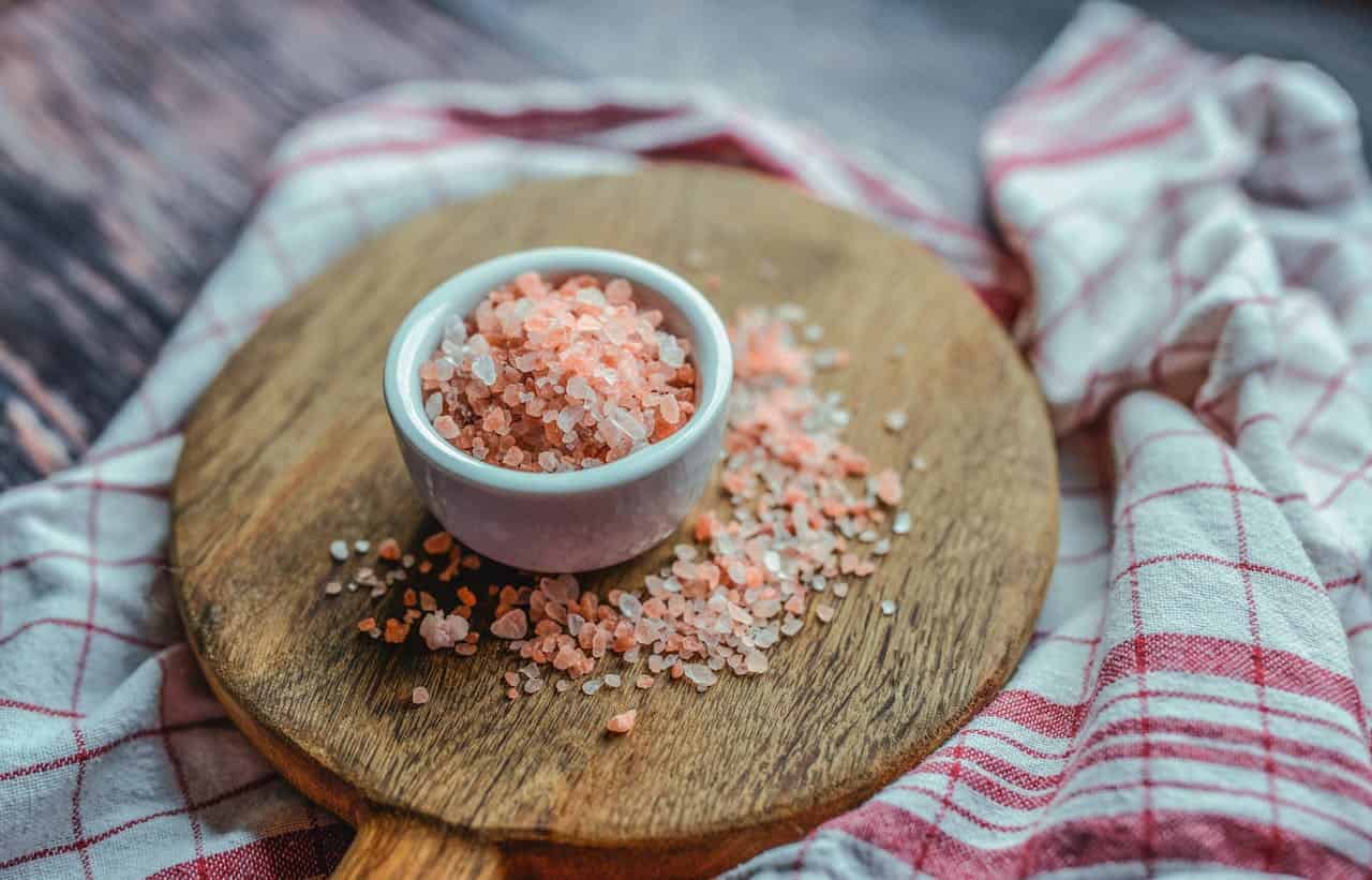 A small white bowl filled with pink Himalayan salt sits on a round wooden cutting board, salt crystals scattered around, a red and white checkered cloth in the background