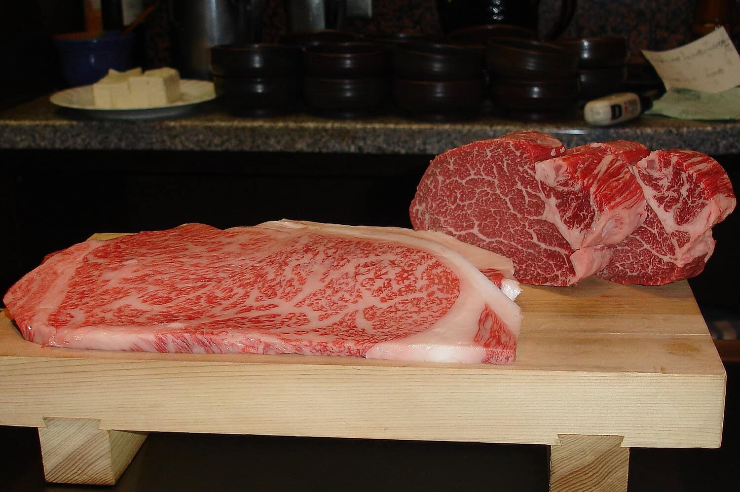 Two cuts of premium marbled beef on wooden cutting board, likely Wagyu or Kobe beef with extensive marbling, kitchen countertop with butter and containers in background