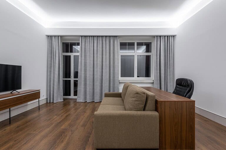 Modern minimalist living room, brown sofa against a wooden desk, black office chair, wooden TV stand with a flat-screen TV, floor-to-ceiling gray curtains, white walls, recessed ceiling lighting, wooden laminate flooring