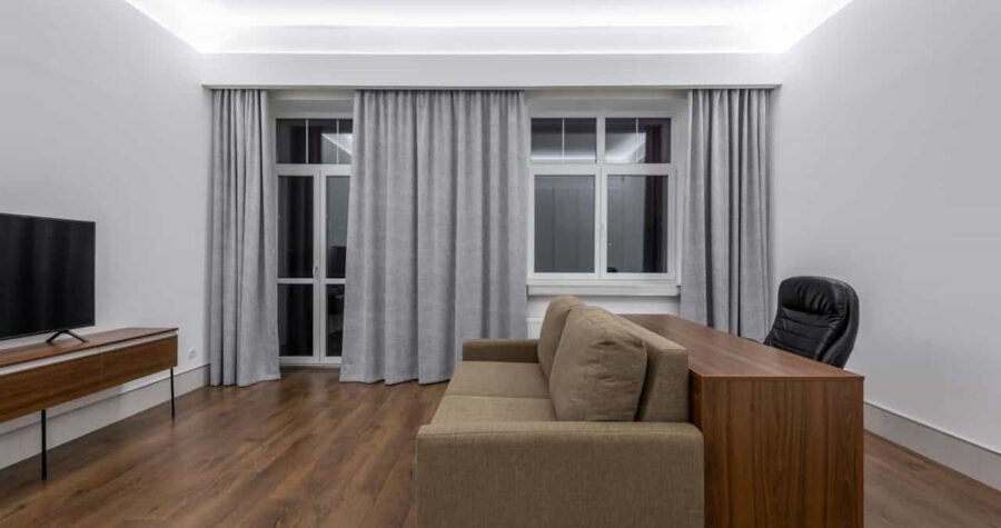 Modern minimalist living room, brown sofa against a wooden desk, black office chair, wooden TV stand with a flat-screen TV, floor-to-ceiling gray curtains, white walls, recessed ceiling lighting, wooden laminate flooring