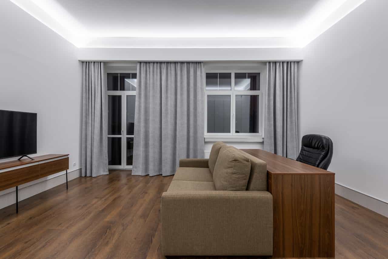 Modern minimalist living room, brown sofa against a wooden desk, black office chair, wooden TV stand with a flat-screen TV, floor-to-ceiling gray curtains, white walls, recessed ceiling lighting, wooden laminate flooring