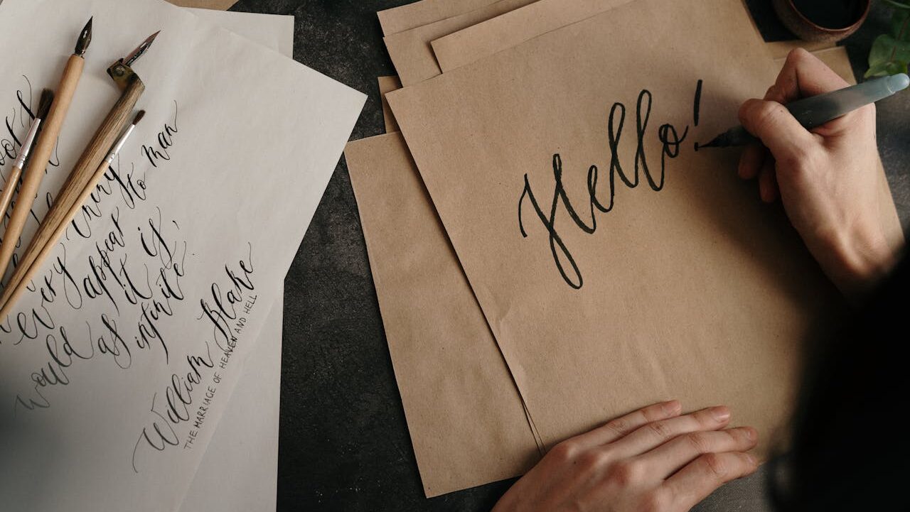 A person writing "Hello!" in elegant calligraphy on brown paper, calligraphy pens and ink on a dark surface, artistic handwriting practice, vintage and creative aesthetic
