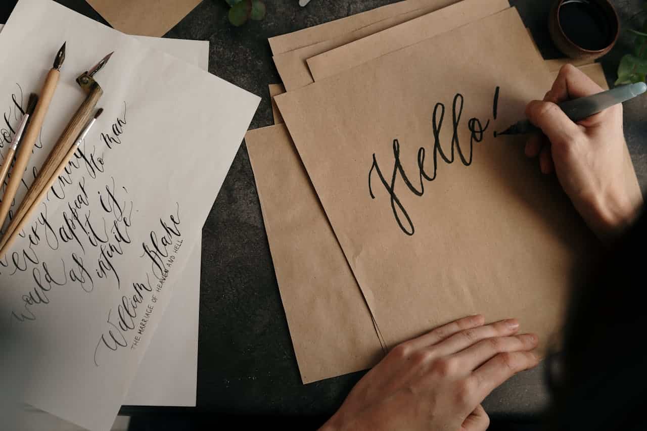 A person writing "Hello!" in elegant calligraphy on brown paper, calligraphy pens and ink on a dark surface, artistic handwriting practice, vintage and creative aesthetic