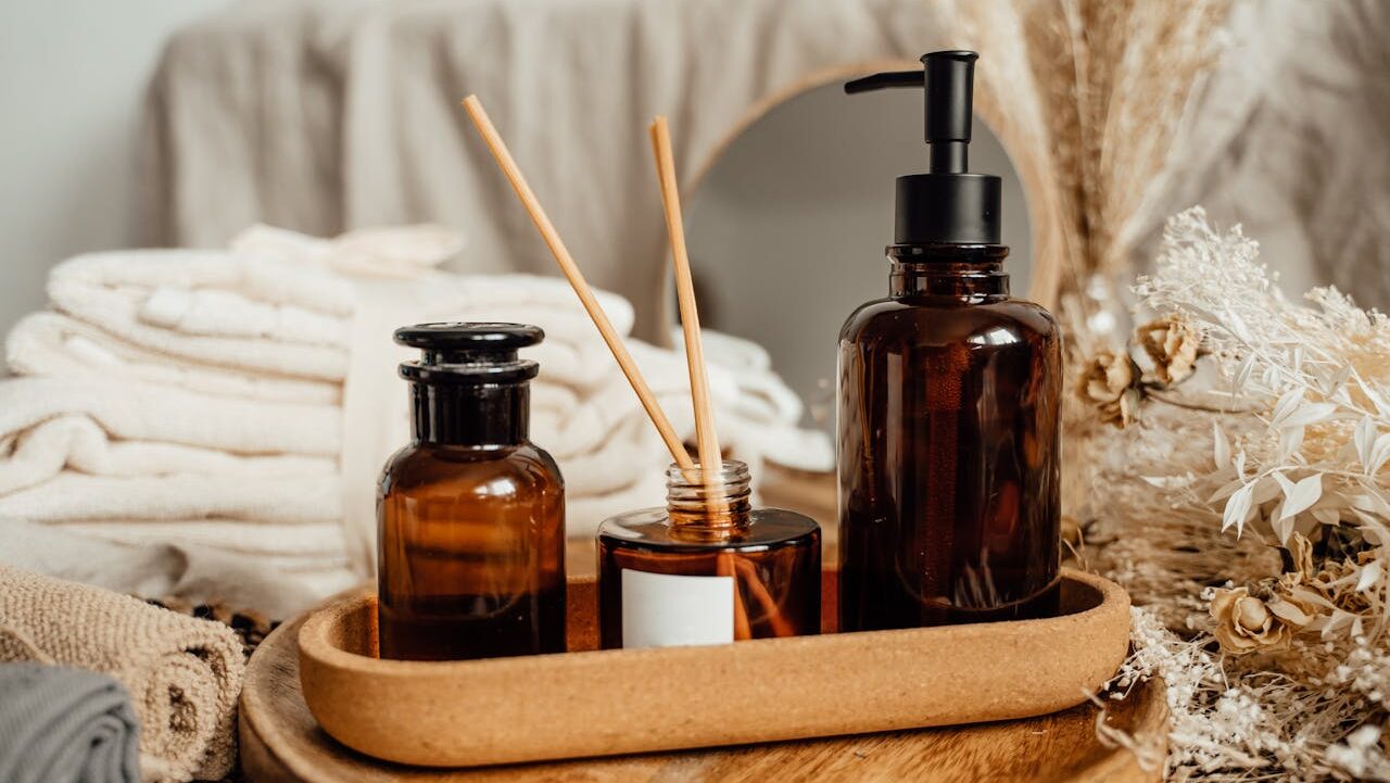 Brown glass bottles with liquid air freshener, wooden diffuser sticks, a soap dispenser, neatly folded towels, a mirror, and dried flowers, arranged on a wooden tray for a spa-like aesthetic