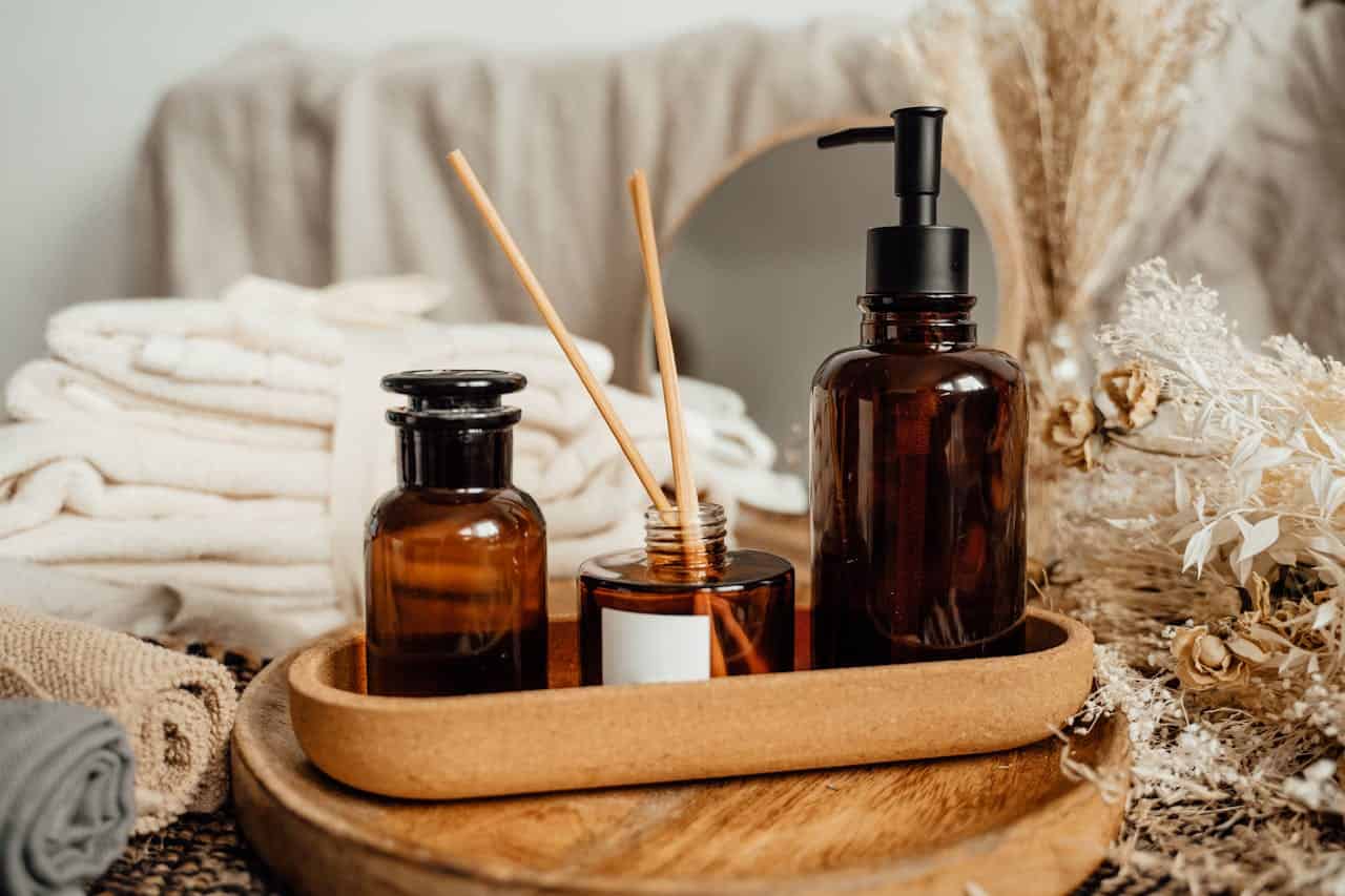 Brown glass bottles with liquid air freshener, wooden diffuser sticks, a soap dispenser, neatly folded towels, a mirror, and dried flowers, arranged on a wooden tray for a spa-like aesthetic