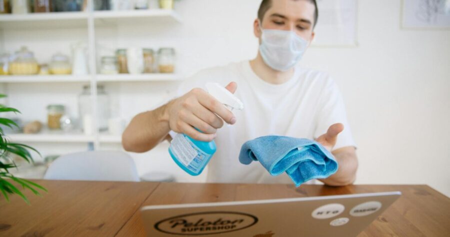 Man using disinfectant to clean laptop with a face mask on.