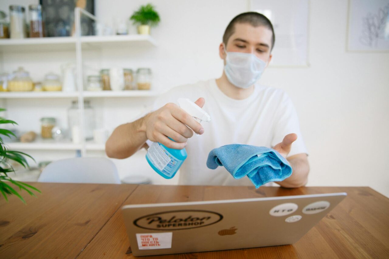 Man using disinfectant to clean laptop with a face mask on.