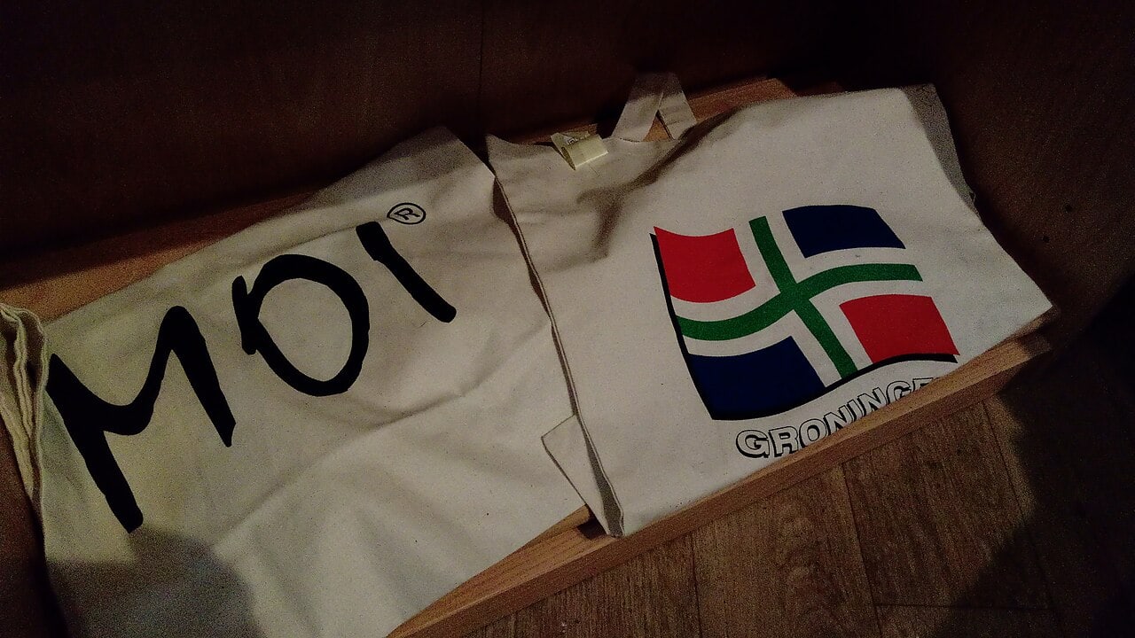 Two white canvas tote bags resting on a wooden surface, one featuring the word "Moi" in black letters, the other displaying a red, blue, and green flag design with the word "Groningen" underneath, both bags are folded with a simple and clean appearance