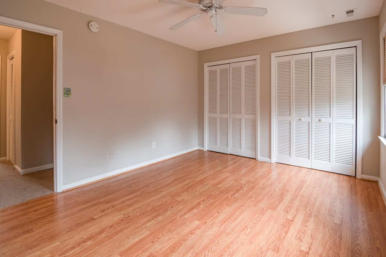 Empty bedroom with light wood flooring, beige walls, white closet doors, a ceiling fan, and natural light from a window, connected to a hallway with carpeted flooring
