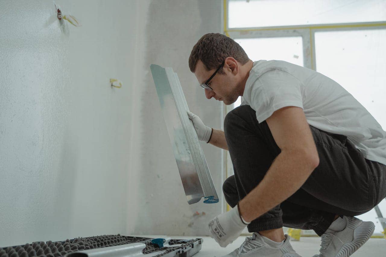 Person in white t-shirt and glasses applying drywall or plaster to wall, wearing work gloves, crouching position, tools visible in foreground, renovation in progress