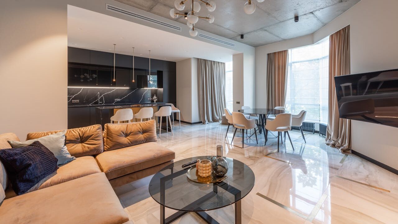 Modern luxury apartment interior with open concept design, beige sectional sofa, glass coffee table, round dining table with white chairs, black marble kitchen, concrete ceiling with gold pendant lights