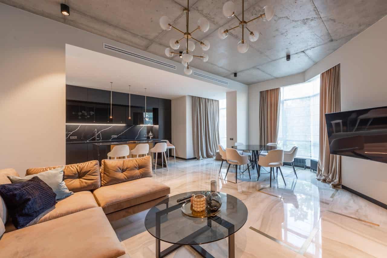 Modern luxury apartment interior with open concept design, beige sectional sofa, glass coffee table, round dining table with white chairs, black marble kitchen, concrete ceiling with gold pendant lights