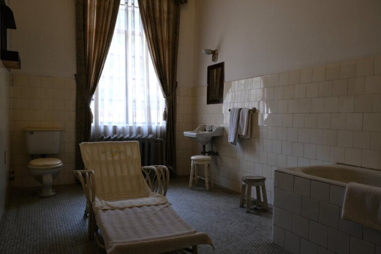 Vintage bathroom, tiled walls, old-fashioned bathtub, classic sink, wooden stools, lounge chair with towel, toilet in the corner, long curtains, nostalgic atmosphere