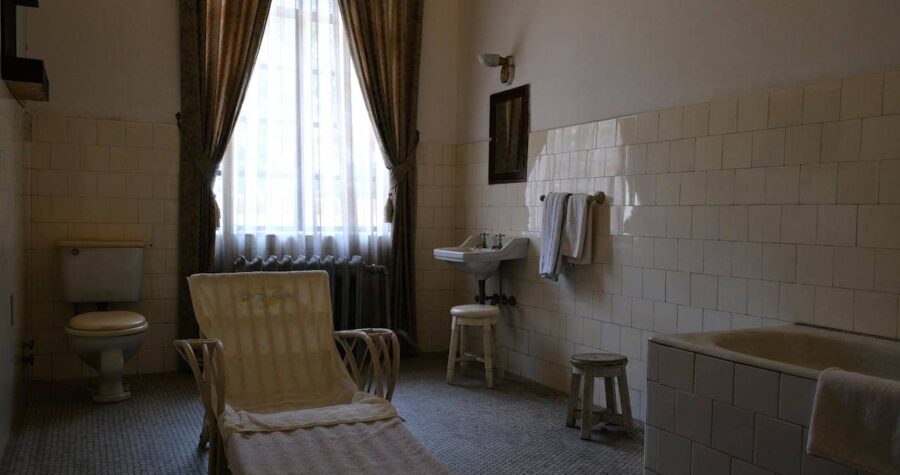 Vintage bathroom, tiled walls, old-fashioned bathtub, classic sink, wooden stools, lounge chair with towel, toilet in the corner, long curtains, nostalgic atmosphere