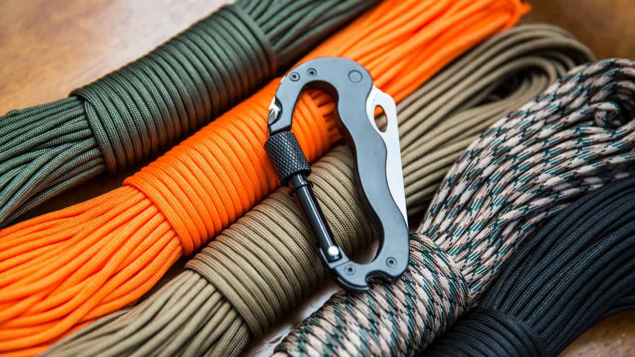 Metal carabiner resting on various colored paracord bundles including green, orange, tan, patterned, and black on wooden surface