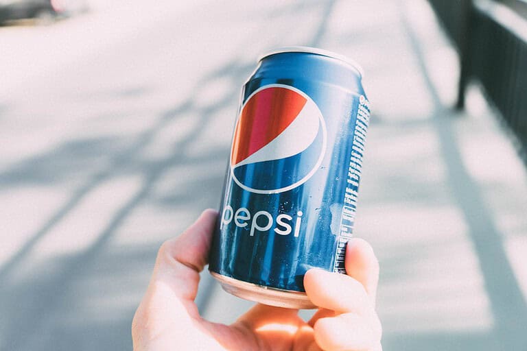 Hand holding a blue Pepsi can, condensation on the surface, blurred outdoor background, bright lighting, carbonated soft drink
