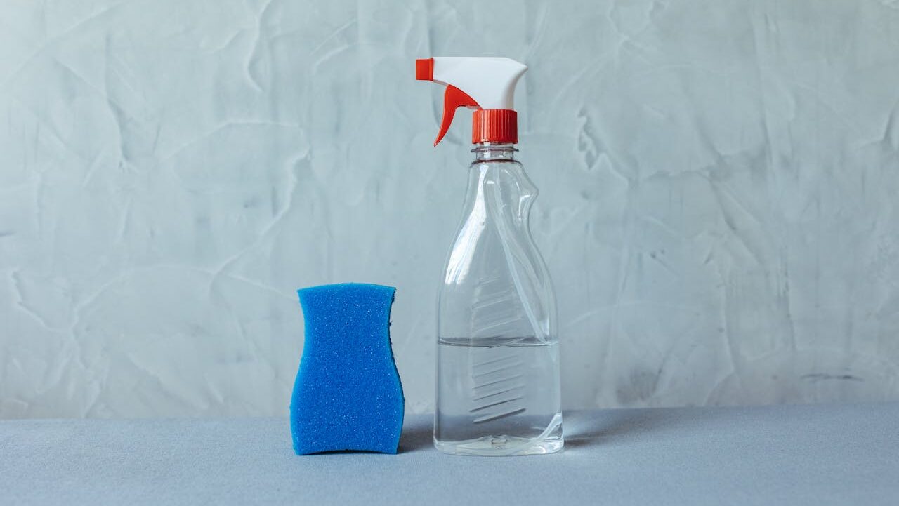 Clear spray bottle with a red nozzle, half-filled with liquid, placed next to a blue cleaning sponge, set against a textured gray wall, minimalistic cleaning setup