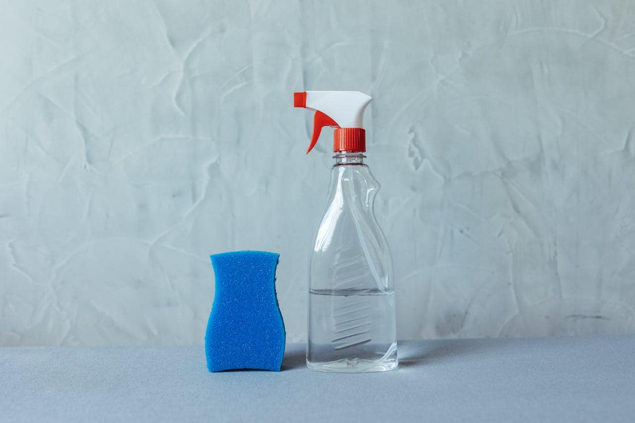 Clear spray bottle with a red nozzle, half-filled with liquid, placed next to a blue cleaning sponge, set against a textured gray wall, minimalistic cleaning setup