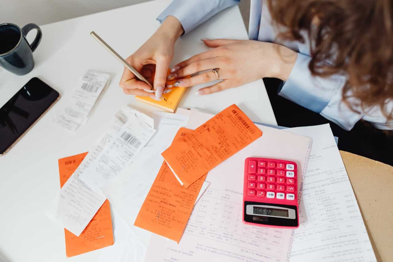 Woman managing finances, writing on a notepad, receipts spread on table, pink calculator, smartphone, coffee cup, financial documents, budgeting, accounting, expense tracking