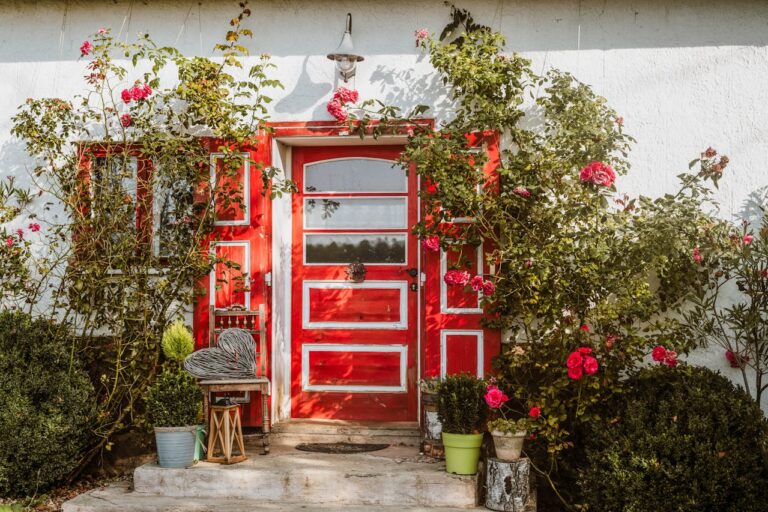 Bright red door with white trim on white house, climbing roses with pink blooms surrounding entrance, decorative lantern and potted plants on small porch, rustic cottage aesthetic
