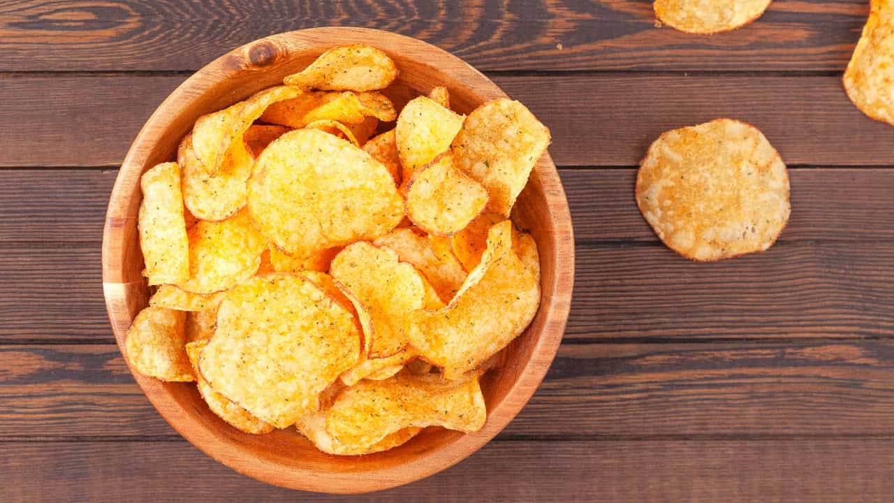 A wooden bowl filled with crispy potato chips, some chips spilling onto the wooden surface, a close-up view of the chips with a golden, textured appearance, the chips are arranged neatly, showing their thin and crunchy texture