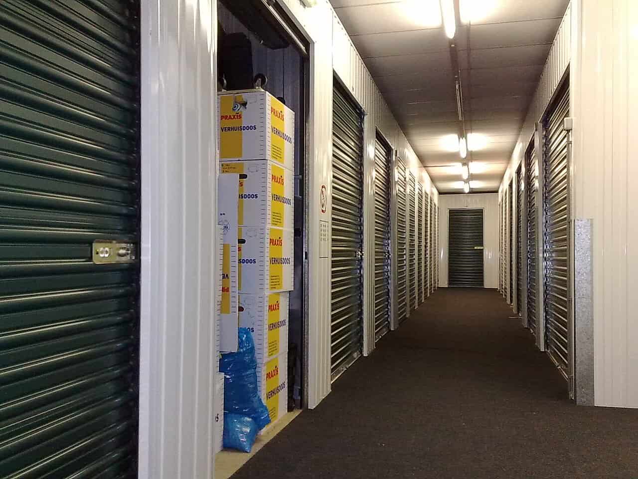 Storage unit hallway with green rolling doors, boxes stacked inside one unit, neat and organized space, fluorescent lighting above, dark carpeted floor, metal panel walls, secured storage area, empty units visible in the background