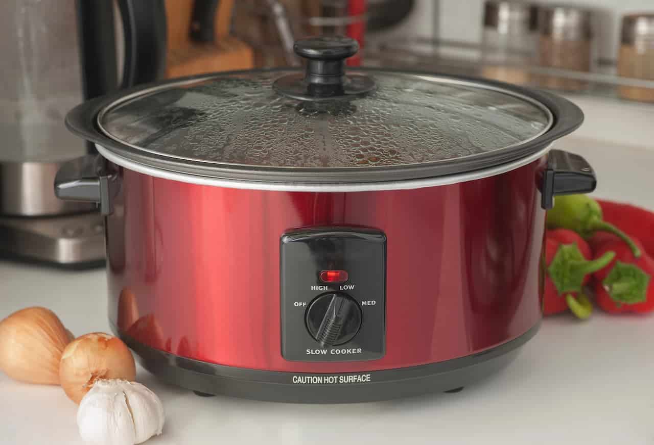 Red slow cooker with a black lid, steam on the glass top, control knob set to medium, onions and garlic on the countertop, fresh red and green peppers in the background, modern kitchen setting