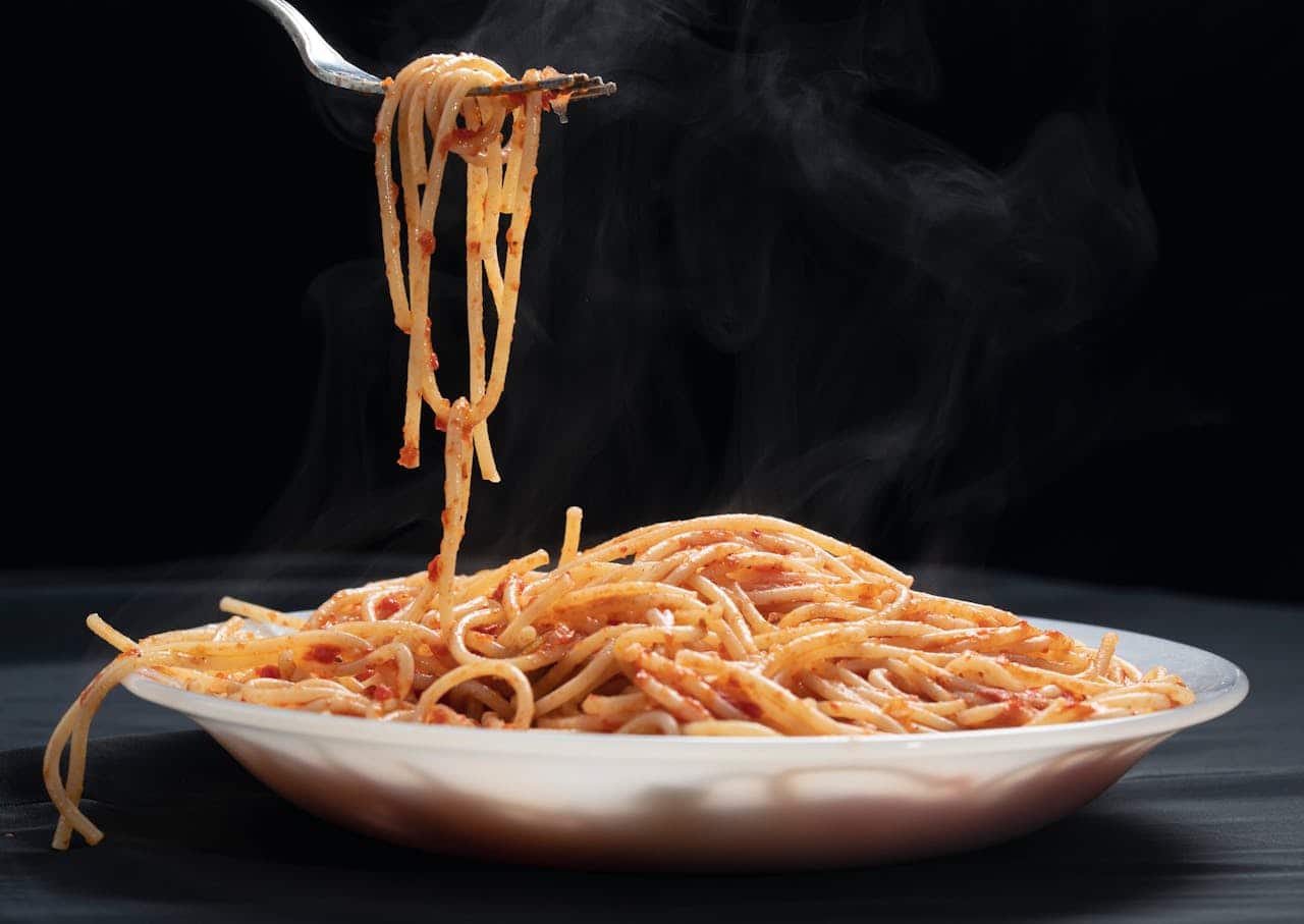 A fork lifting steaming spaghetti with tomato sauce from a white plate, dark background, rising steam, pasta coated in red sauce
