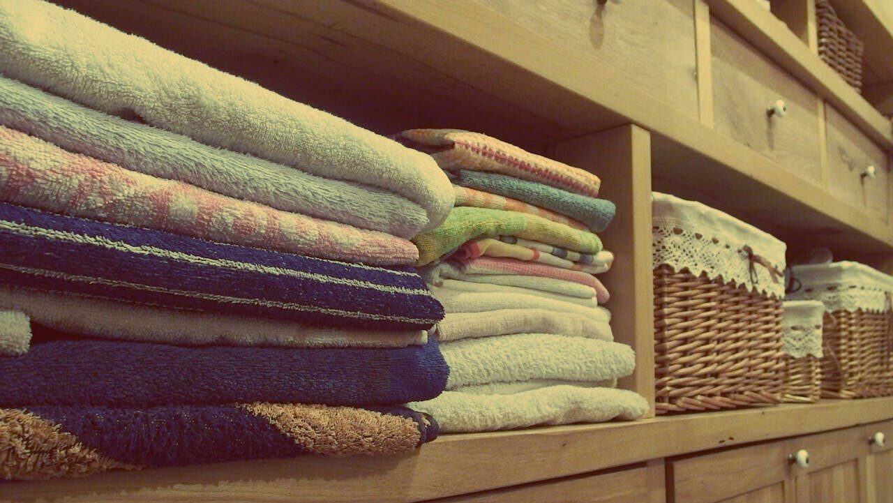 Neatly folded towels stacked on a wooden shelf, colorful and soft, next to woven wicker baskets with lace covers, part of an organized linen closet or bathroom storage