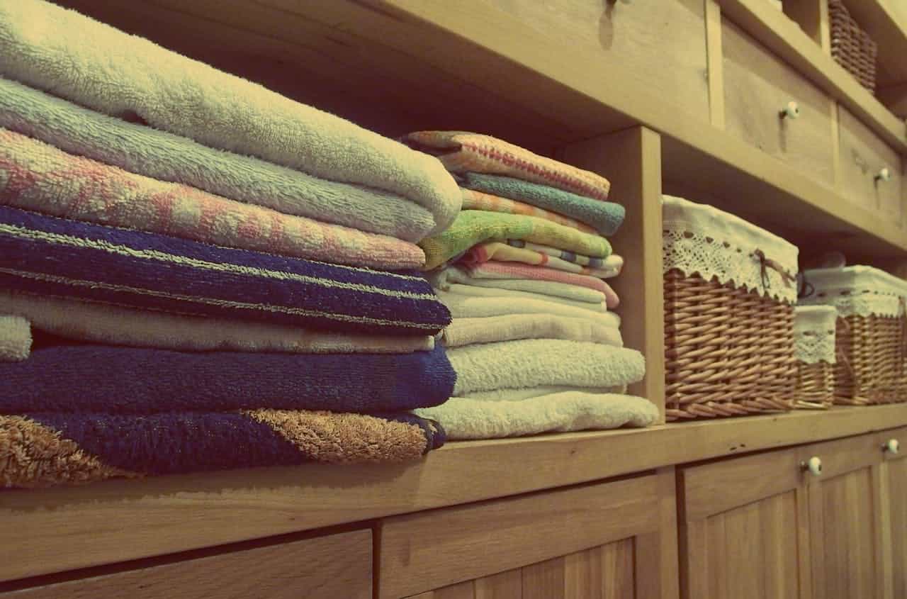 Neatly folded towels stacked on a wooden shelf, colorful and soft, next to woven wicker baskets with lace covers, part of an organized linen closet or bathroom storage