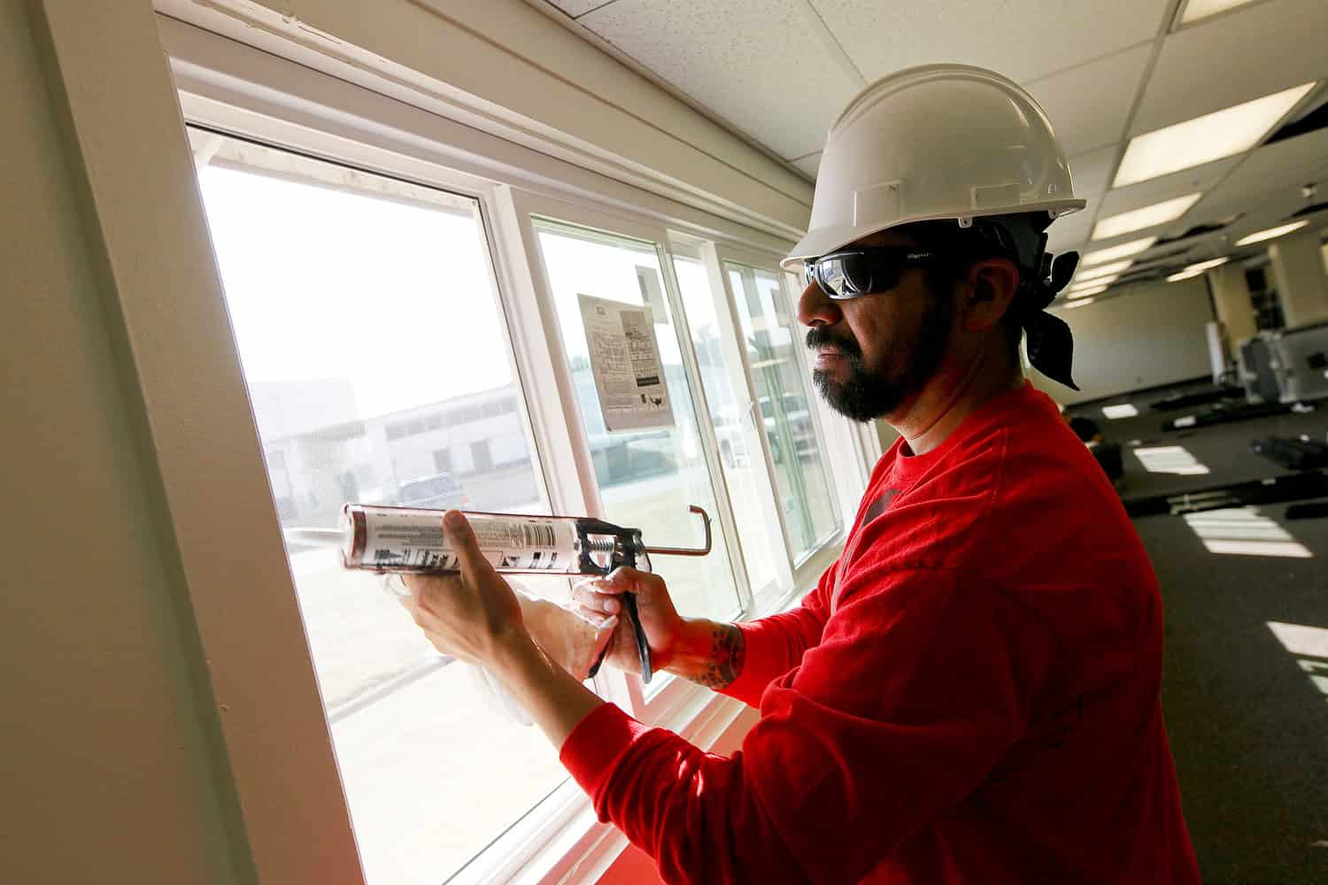 A construction worker applying caulk to a window frame, wearing a white hard hat, sunglasses, and a red shirt, focused on sealing the window