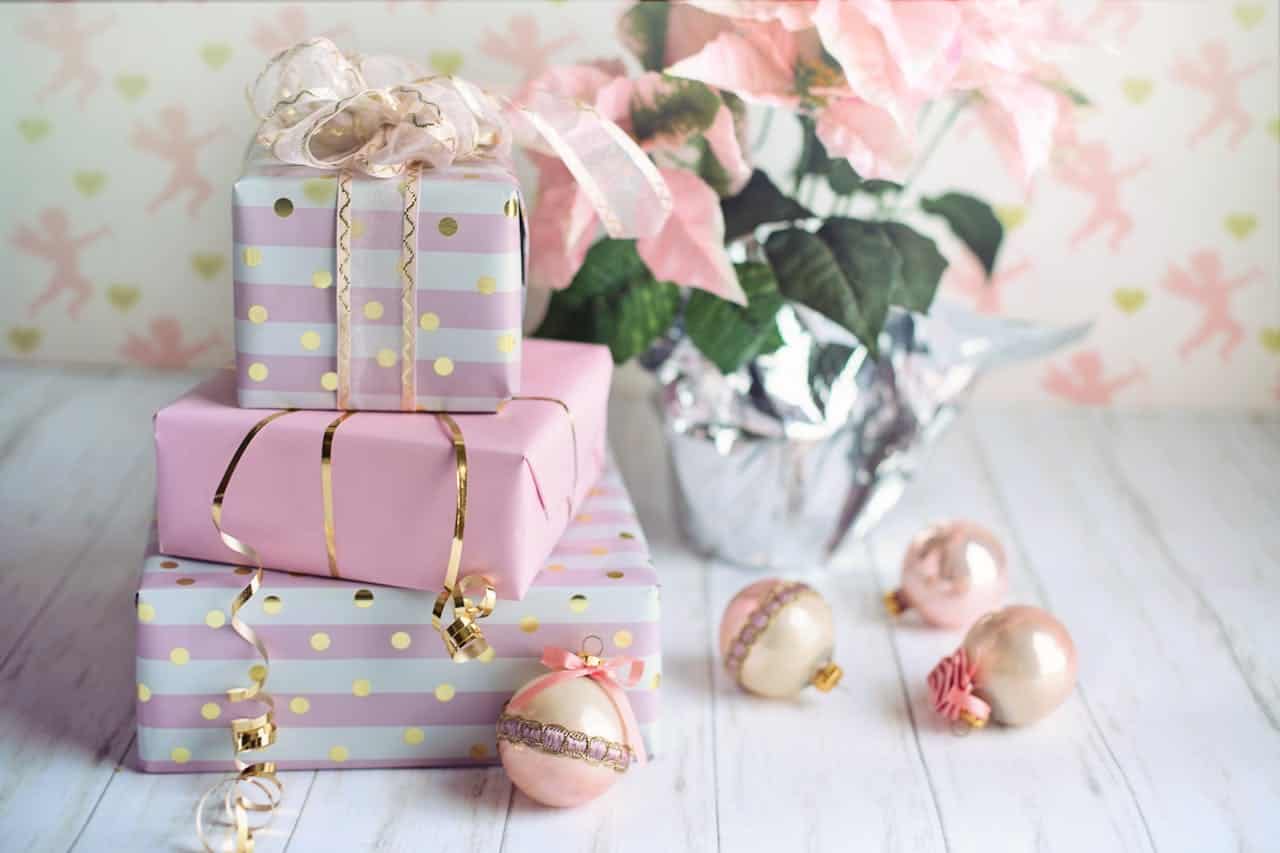 Stacked pink and gold-wrapped gifts with ribbons, delicate pink and gold Christmas ornaments, a pink poinsettia in a foil-wrapped pot, soft pastel background with cherub and heart patterns, white wooden surface