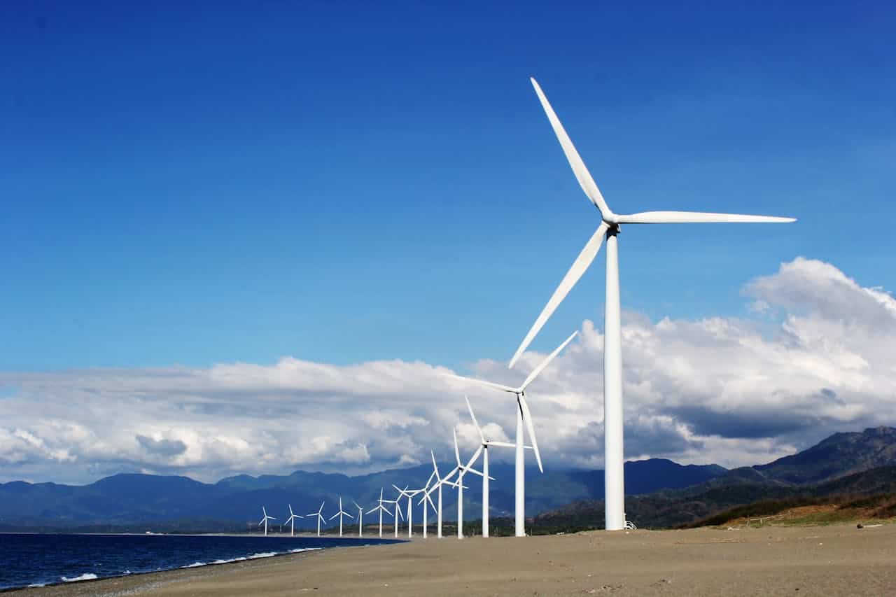 White wind turbines lined along a sandy coastline, blue sky with clouds, mountain range in background, shoreline visible, renewable energy installation