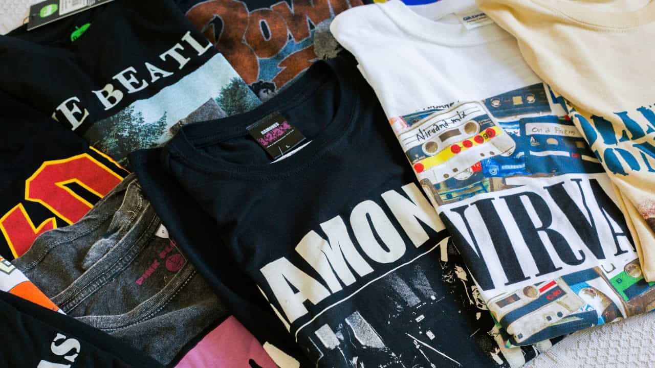 Black t-shirt with "AMONOS" text, laid on colorful fabric or clothing items, partial view of tags, blue checkmark icon in upper right corner