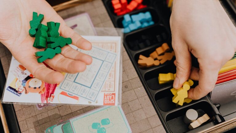Close-up of two hands handling colorful wooden board game pieces, one holding green meeples, the other picking a yellow meeple from a black organizer tray filled with sorted game components, board and cards partially visible