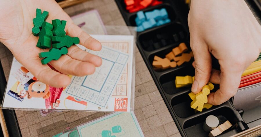 Close-up of two hands handling colorful wooden board game pieces, one holding green meeples, the other picking a yellow meeple from a black organizer tray filled with sorted game components, board and cards partially visible