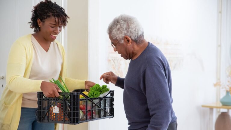 A smiling woman in a yellow cardigan hands a crate of fresh vegetables to an elderly man in a navy sweater, indoor setting, act of kindness, food delivery, grocery assistance