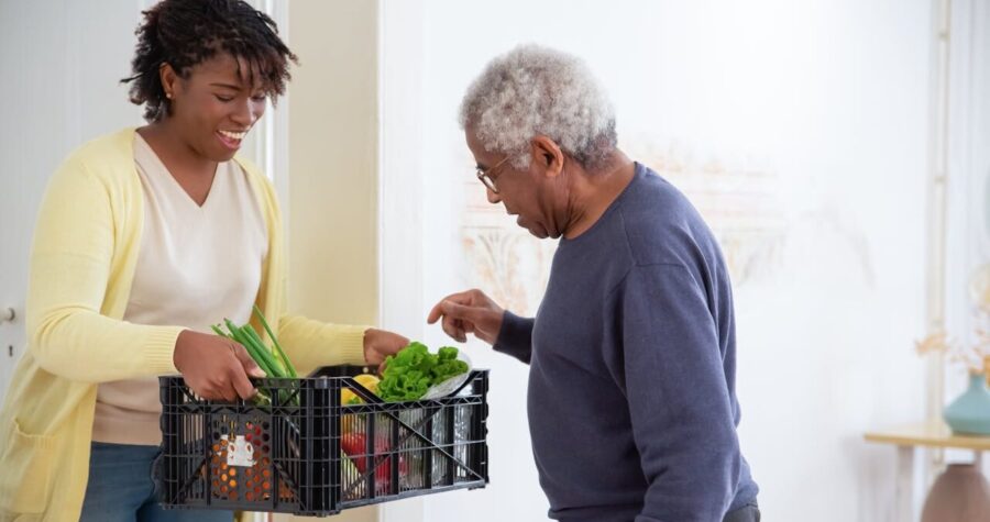 A smiling woman in a yellow cardigan hands a crate of fresh vegetables to an elderly man in a navy sweater, indoor setting, act of kindness, food delivery, grocery assistance