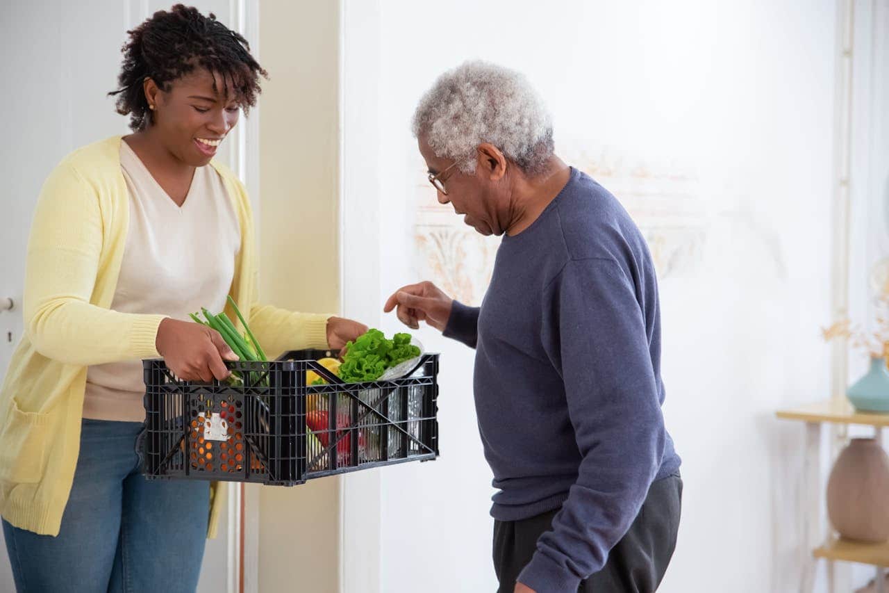 A smiling woman in a yellow cardigan hands a crate of fresh vegetables to an elderly man in a navy sweater, indoor setting, act of kindness, food delivery, grocery assistance