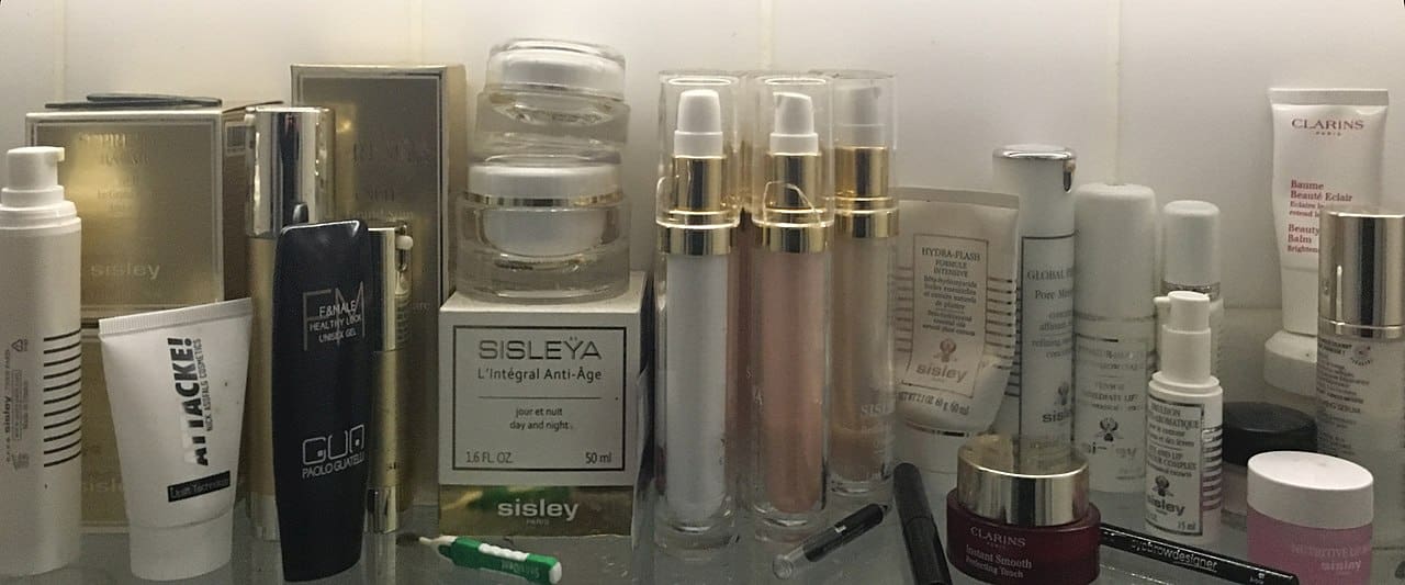 Collection of luxury skincare products, Sisley and Clarins brands, various bottles and tubes, crowded bathroom shelf, anti-aging creams and serums