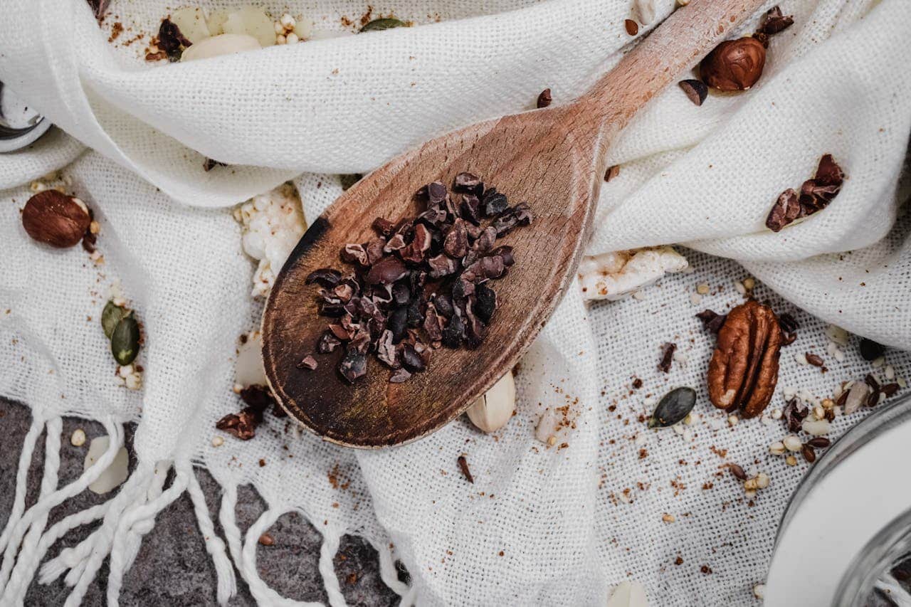 Wooden spoon with cacao nibs, scattered nuts and seeds, textured white fabric, rustic and organic food setting, earthy tones