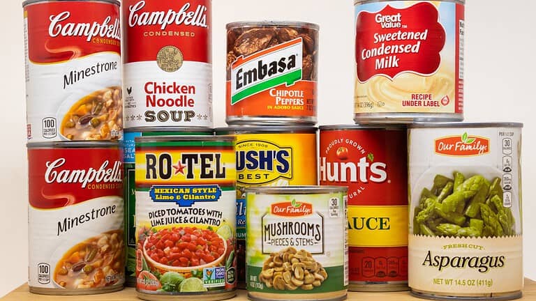 Stack of assorted canned foods including soups, tomatoes, beans, condensed milk, mushrooms, sauce, and asparagus