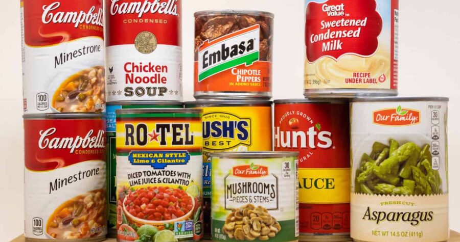 Stack of assorted canned foods including soups, tomatoes, beans, condensed milk, mushrooms, sauce, and asparagus