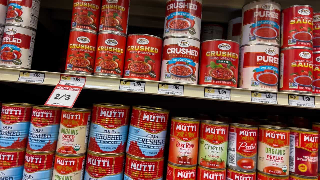 Shelves stocked with canned crushed tomatoes, various brands with red and blue labels, price tags visible, grocery store setting, products arranged for sale, commonly used for cooking, meal preparation, and pantry stocking